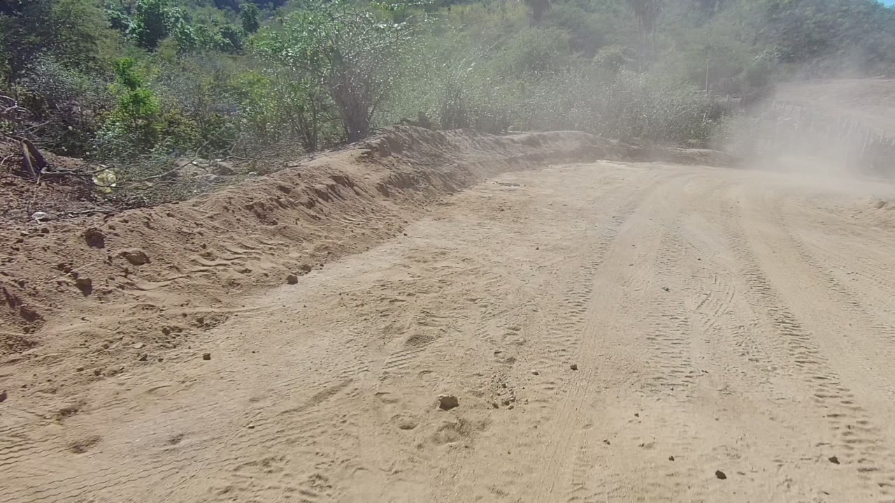 The road extends forward through dry Los Cabos terrain, with tire tracks in the foreground, dust drifting across the midground, and shrubs along both sides, with no visible people.