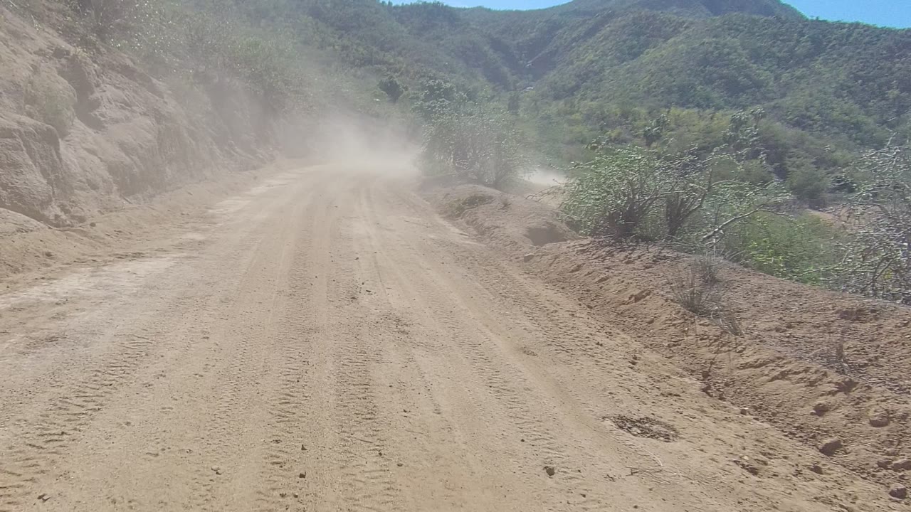 A dusty unpaved road in Los Cabos, Baja California Sur runs between rocky hillsides and green shrubs, with no visible people.