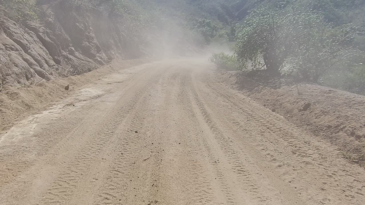 A dust-covered road in Los Cabos passes a rocky slope on the right and sparse shrubs on both sides, with no visible people.