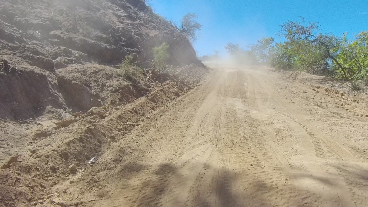 A dusty dirt road in Los Cabos, Baja California Sur rises between a rough rocky bank and sparse green trees under bright blue sky, with no visible people.
