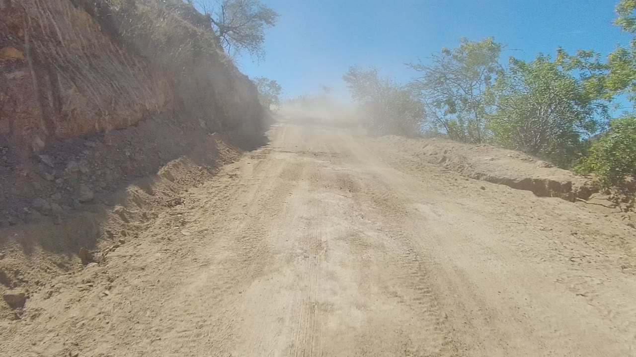 A pale dirt road in Los Cabos, Baja California Sur leads toward a dust cloud, with a steep rock face on the left, green brush on the right, and no visible people.