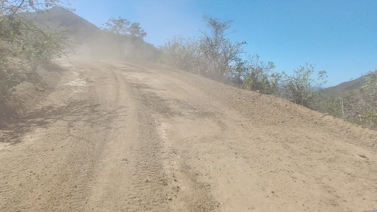 A dry dirt road in Los Cabos, Baja California Sur climbs toward low hills under a clear blue sky, with tire tracks and no visible people.