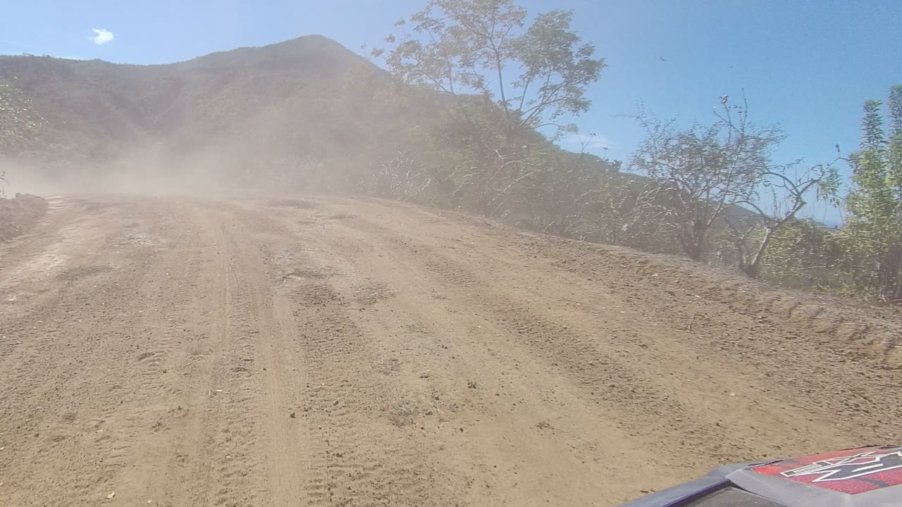 A dusty Los Cabos dirt road curves through dry brush and hills, with part of an off-road vehicle visible at the lower right and no visible people.