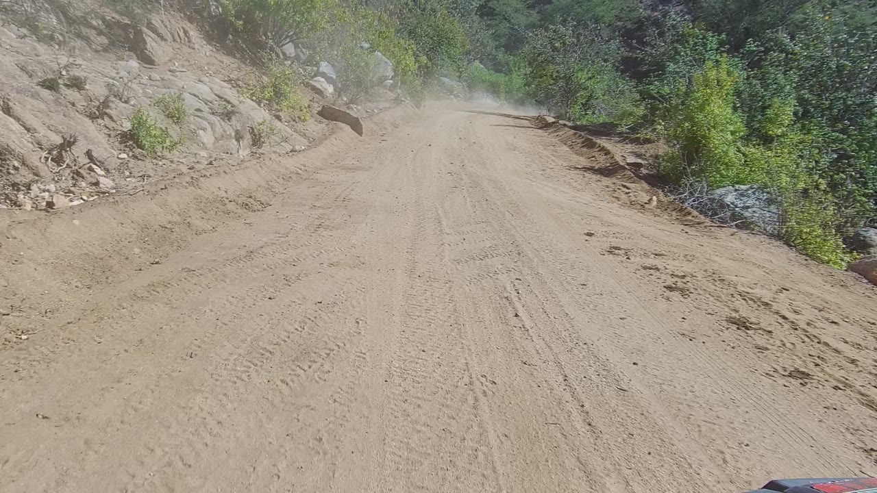 A sandy mountain dirt road in Los Cabos curves ahead between rocky slopes and green roadside brush, with dust visible in the distance and no visible people.