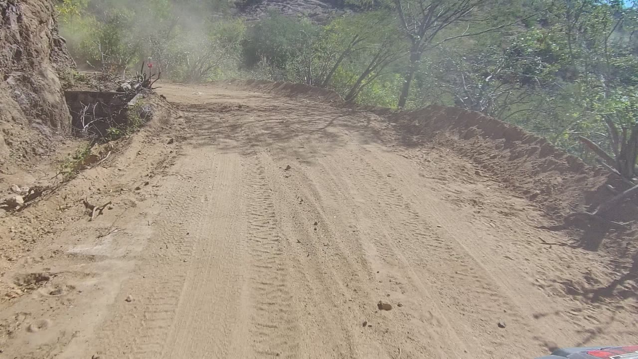 Forward-facing vehicle view of a dusty dirt road in Los Cabos, Mexico, with tire tracks, scrub vegetation, rocky slope, and no visible people.