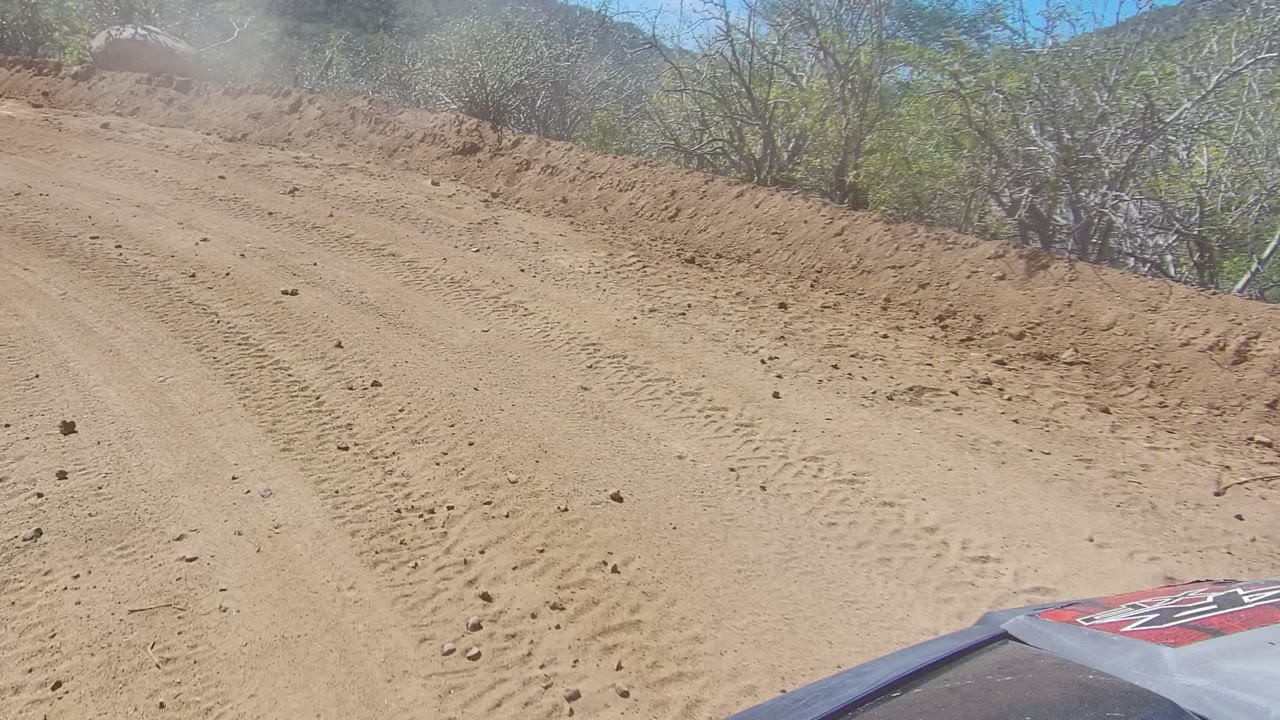 Dust hangs over a sandy unpaved road in Los Cabos, Baja California Sur, with tire tracks, low brush, distant hills, and no visible people.