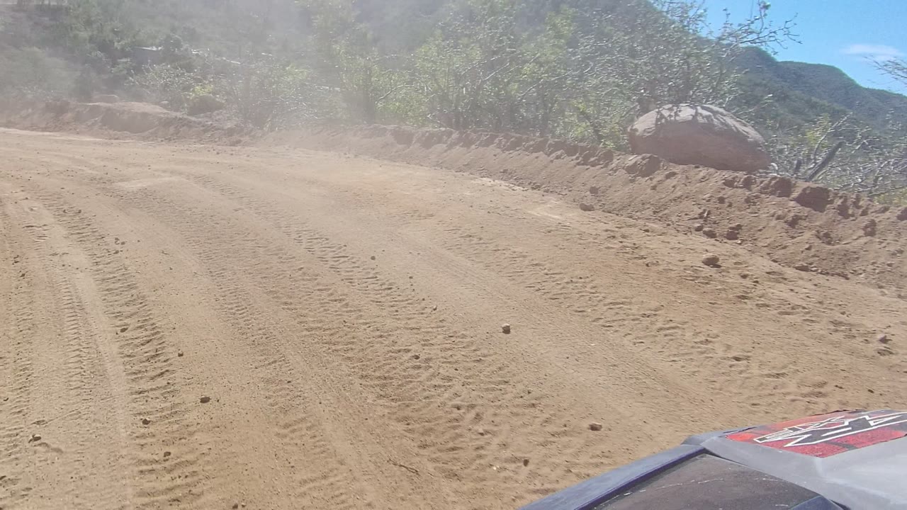 A low vehicle POV follows a dusty dirt road bend in Los Cabos, Baja California Sur, with rocky banks, sparse trees, and no visible people.