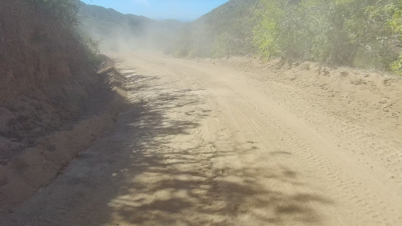 Sandy dirt road in Los Cabos, Baja California Sur, seen from a vehicle with scrub vegetation, rocky hills, bright sun, and no visible people.