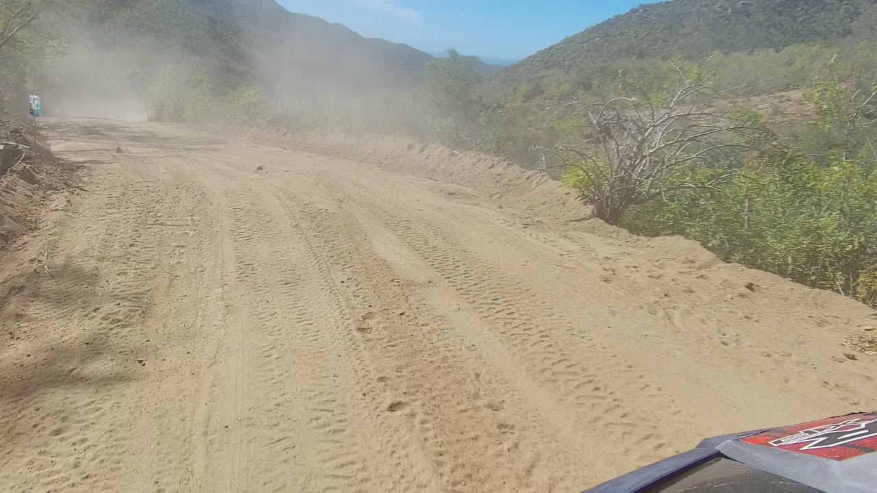 A portion of the vehicle hood appears at the lower edge while the Los Cabos dirt road rises ahead through dust, brush, and dry mountain terrain with no visible people.