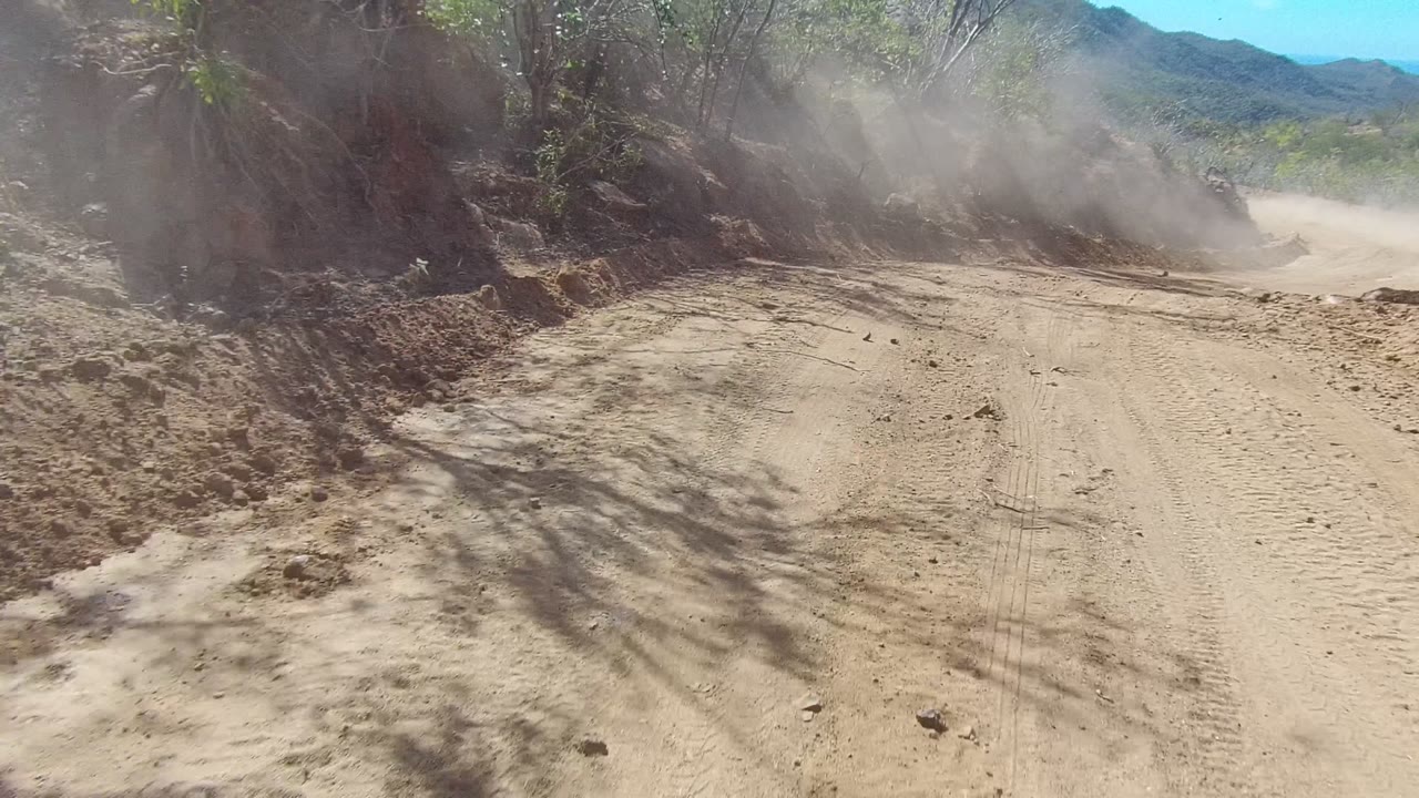 Unpaved road with dust hanging beside a rocky cut bank.