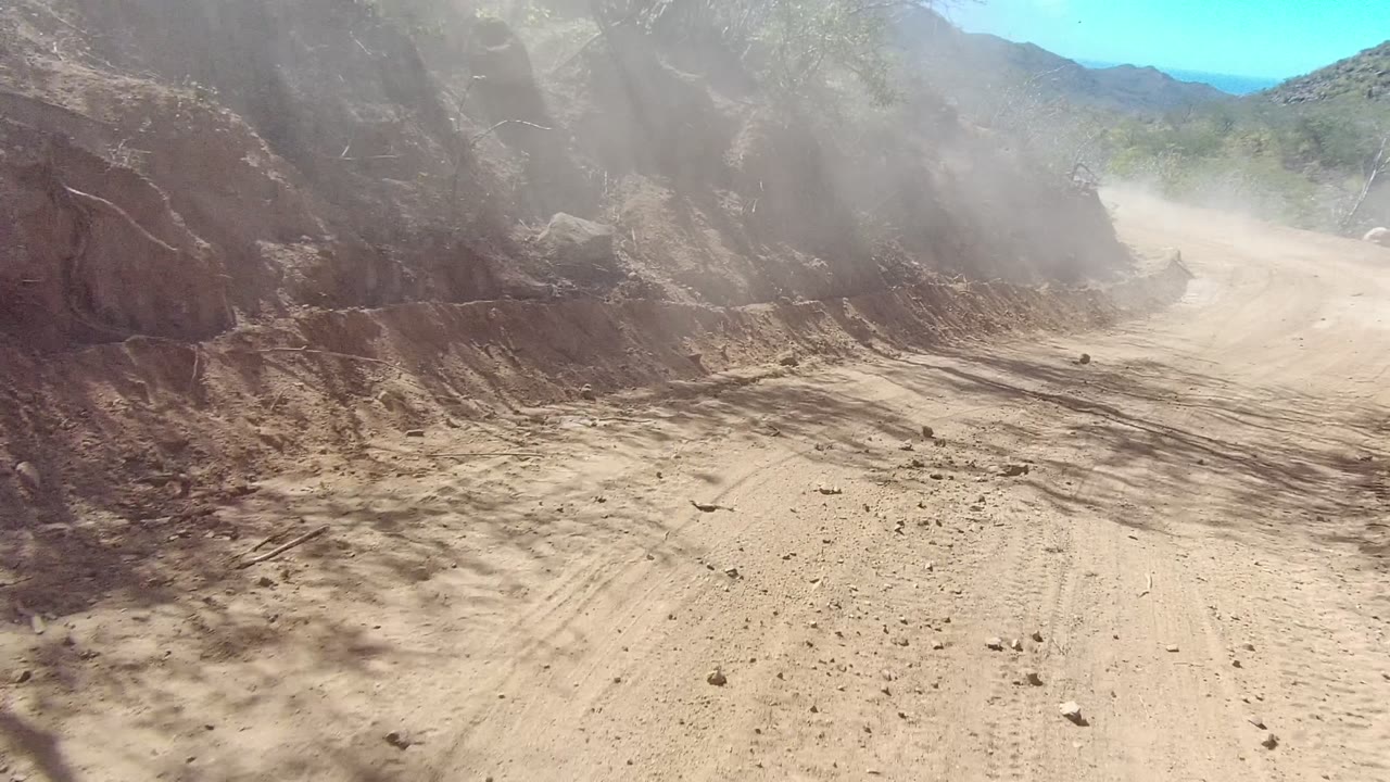 A rough Los Cabos dirt road bends right beside a high rocky bank, with loose stones on the road and dust softening the bright mountain background, with no visible people.