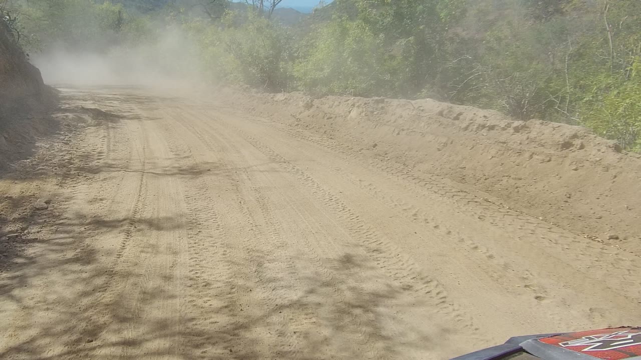 Forward view along a dusty dirt road bordered by brush and hillside cuts.