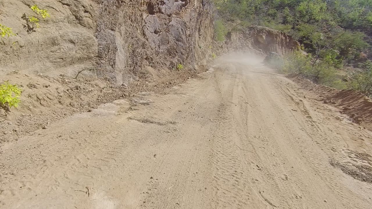 A narrow dirt road in Los Cabos, Baja California Sur runs between rough brown rock walls, with tire tracks in the foreground, sparse greenery at the edges, dust ahead, and no visible people.