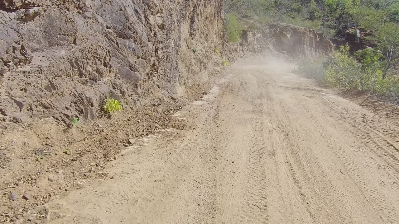 Dusty unpaved road curving through rock cuts and brush.