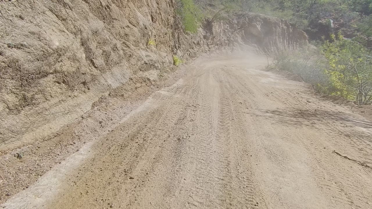 A rutted dirt track in Los Cabos, Baja California Sur fills the foreground, bordered by eroded tan rock on the right, scattered shrubs on the left, dusty air ahead, and no visible people.