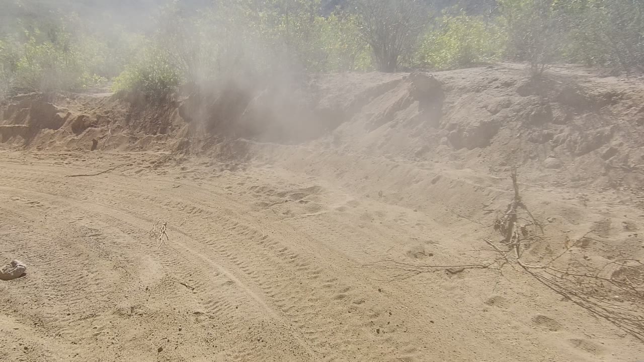 Opening view of a sandy dirt road in Los Cabos with tire tracks, dust hanging over the left side, scrubby bushes on the slopes, and no visible people.