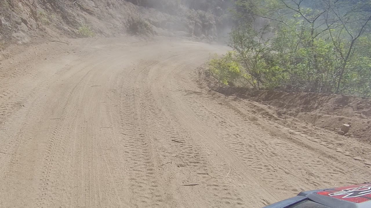 A dusty dirt road in Los Cabos, Baja California Sur curves through rocky terrain with tire tracks, scattered green shrubs, a sliver of vehicle body at the lower edge, and no visible people.