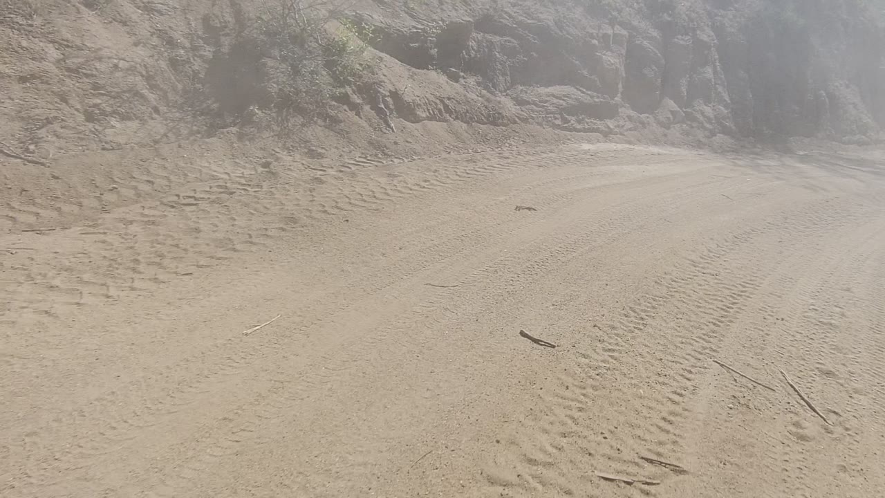 A close road-level view in Los Cabos, Baja California Sur shows dusty tire tracks, a small branch on the dirt, a rock wall at the side, and no visible people.