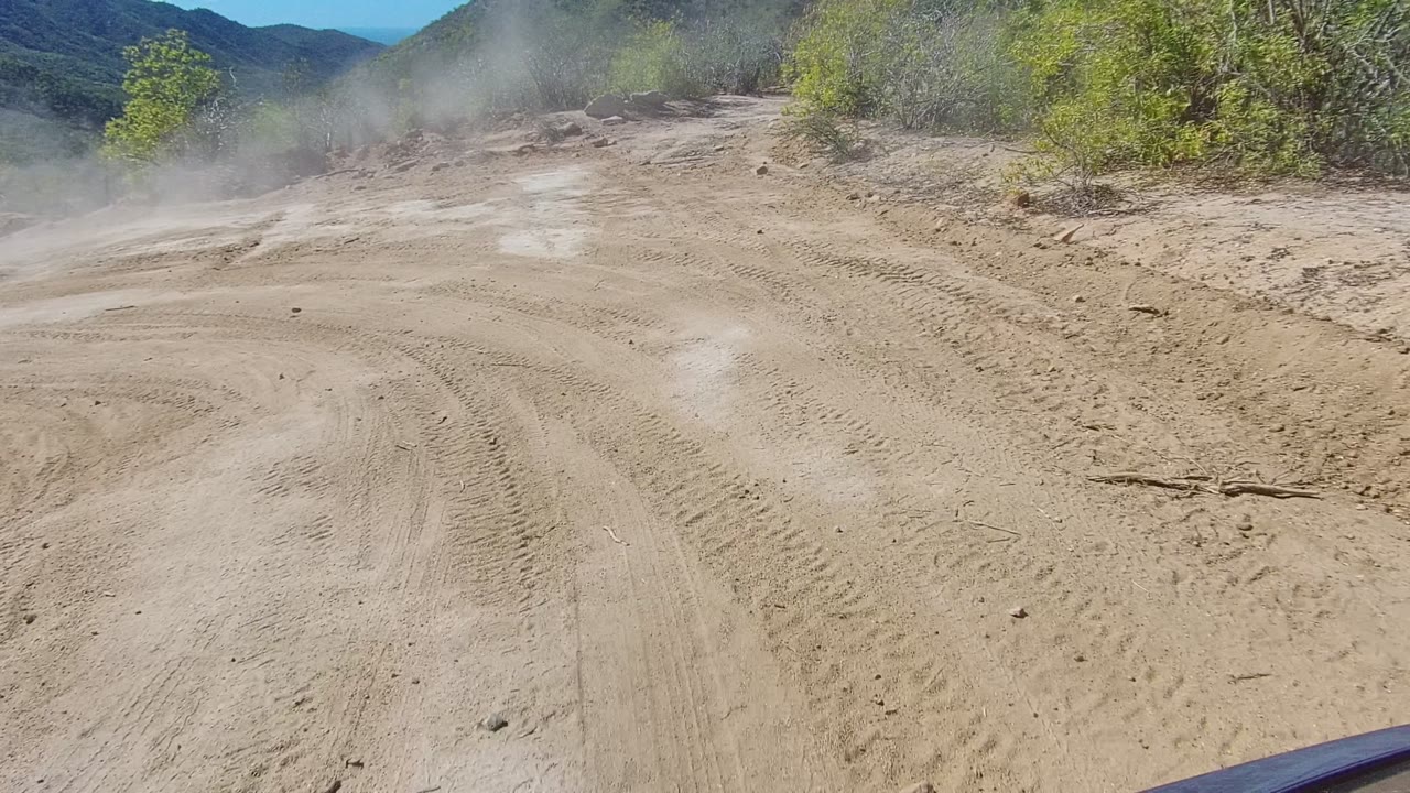 Opening view of a dusty dirt road in Los Cabos, Baja California Sur, with tire tracks, low brush, distant hills, part of the vehicle hood, and no visible people.