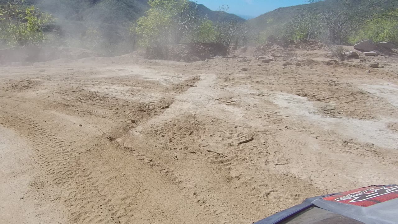 A wider section of dusty Los Cabos dirt road shows rutted sand, pale dust near the brush line, distant hilly terrain, the vehicle hood edge, and no visible people.