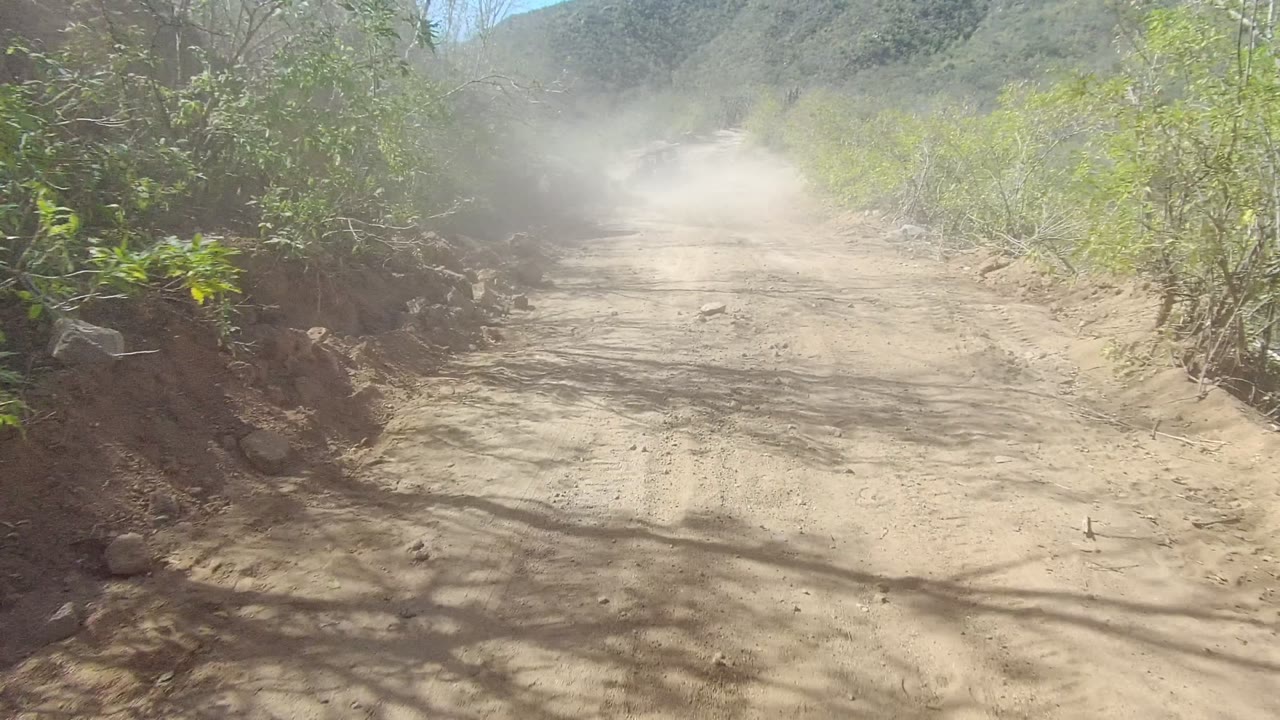 A broader bend in the Los Cabos dirt road opens to distant hills, with dust drifting across the track, green brush at the edges, rocks near the shoulder, and no visible people.