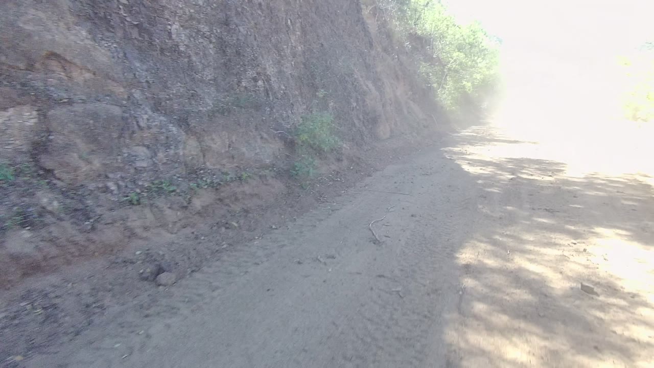 A dusty dirt road in Los Cabos, Baja California Sur runs beside a steep rocky cut bank with green brush on the opposite side and no visible people.