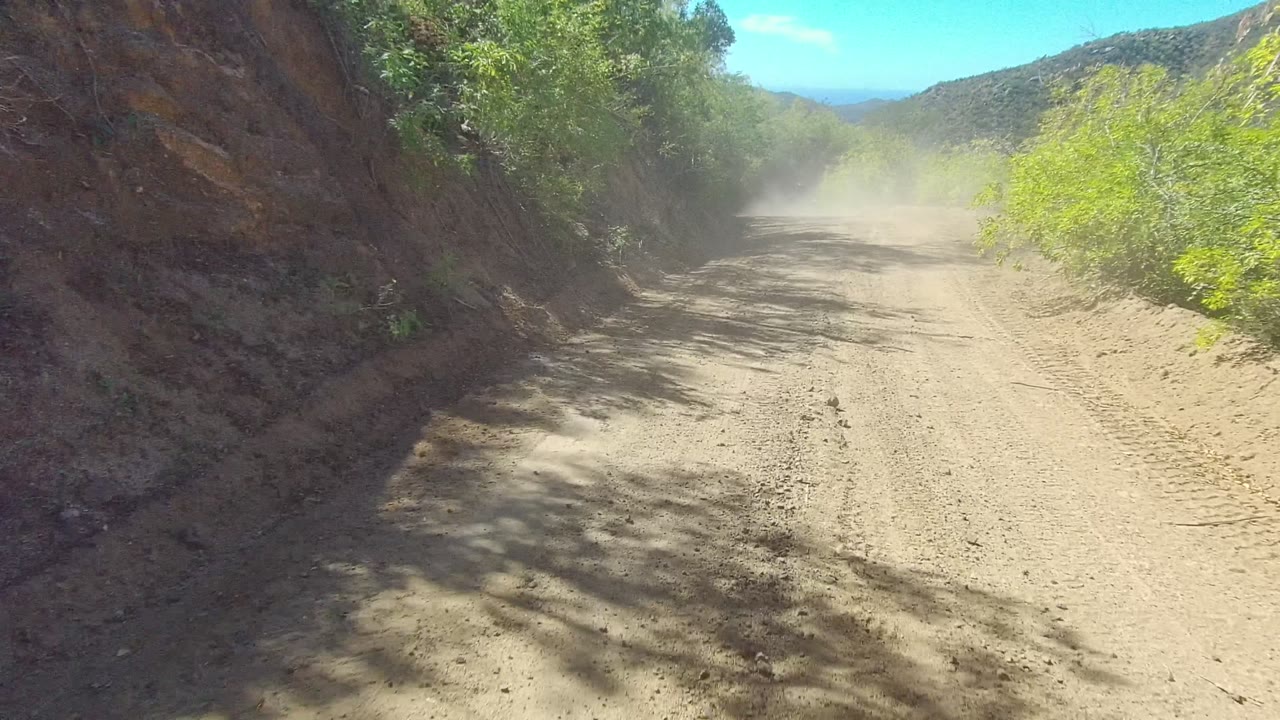 A dusty off-road trail in Los Cabos stretches past the rocky left bank toward scrub-covered hills, with no visible people and light haze over the road.
