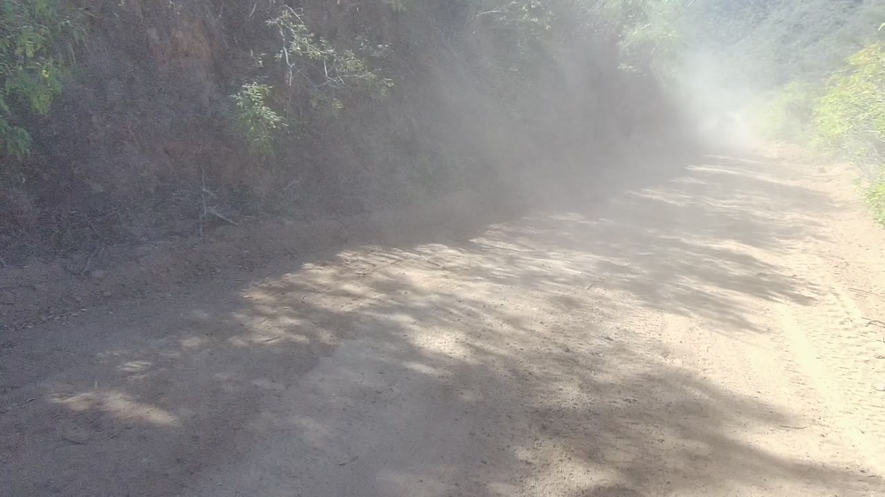 A pale dust cloud drifts over a sunlit dirt road bend in Los Cabos, with dark rock and scrub vegetation framing the road and no visible people.