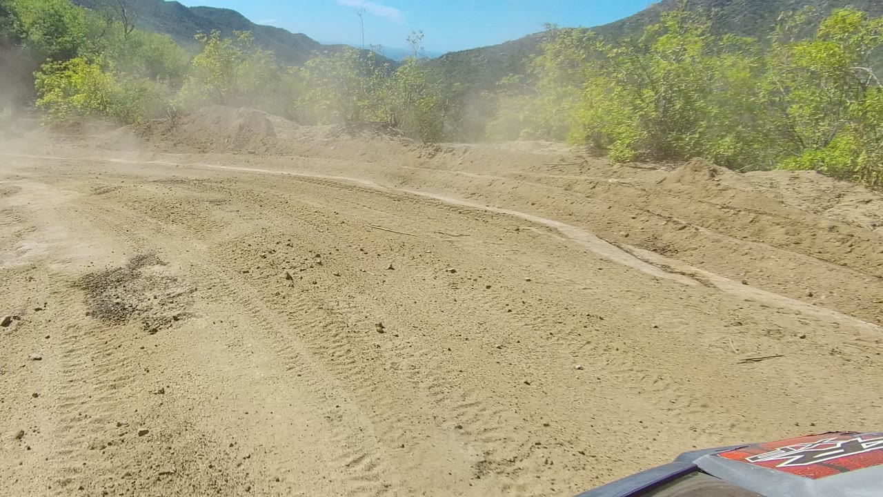 Sandy mountain road curving past brush with dust hanging over the track.