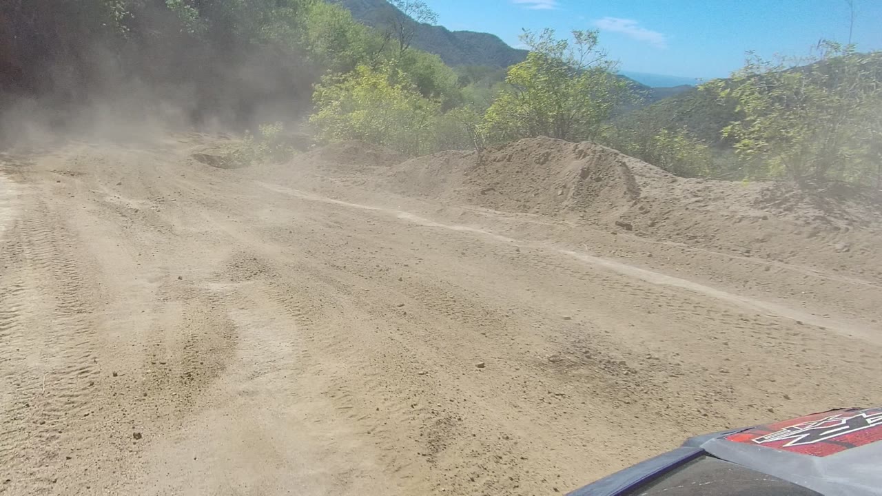 A darker dust plume rises ahead on the Los Cabos dirt road, with sandy tracks in the foreground, brush along both sides, mountains beyond, and no visible people.