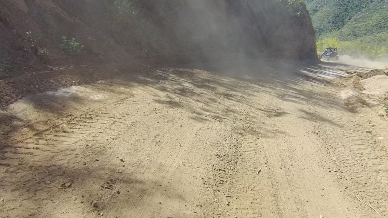 Dust cloud over a dirt road bend between rocky slopes.