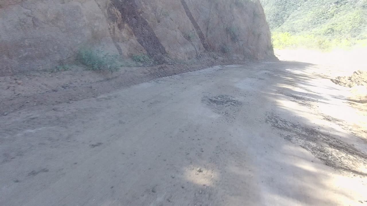 The dirt road runs alongside a tall rock face in Los Cabos, with a pale dusty surface, bright sunlight at the far bend, and no visible people.
