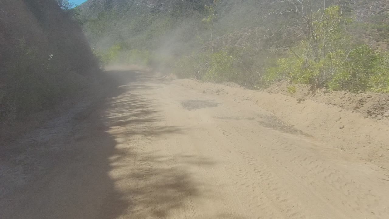 Early frame of a dusty dirt road in Los Cabos, Baja California Sur, with a shaded rocky slope on the left, green brush on the right, and no visible people.