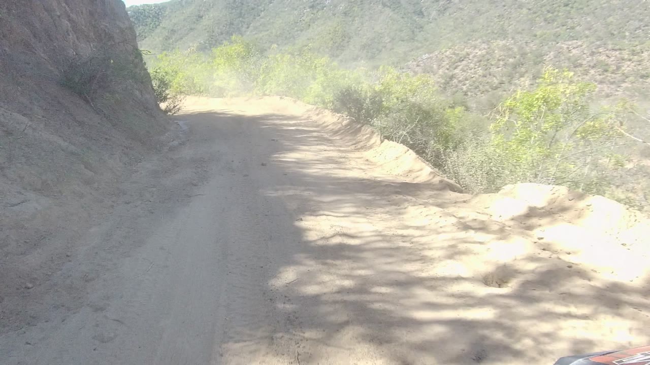 Mid-clip view of a pale dirt road in Los Cabos, Baja California Sur, bending past a rock face with green brush and hilly background terrain, with no visible people.