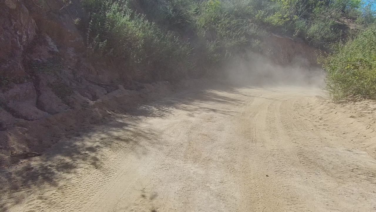 Dust hangs ahead on a winding dirt road in Los Cabos, Baja California Sur, with rocky banks, green brush, tire marks, and no visible people.