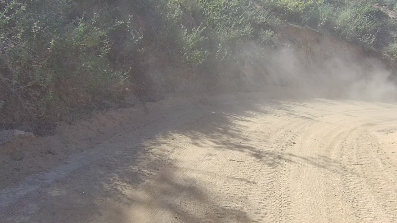 A tire-marked dirt road curves right through Los Cabos brush and rocky terrain as a dust plume drifts across the sunlit road, with no visible people.