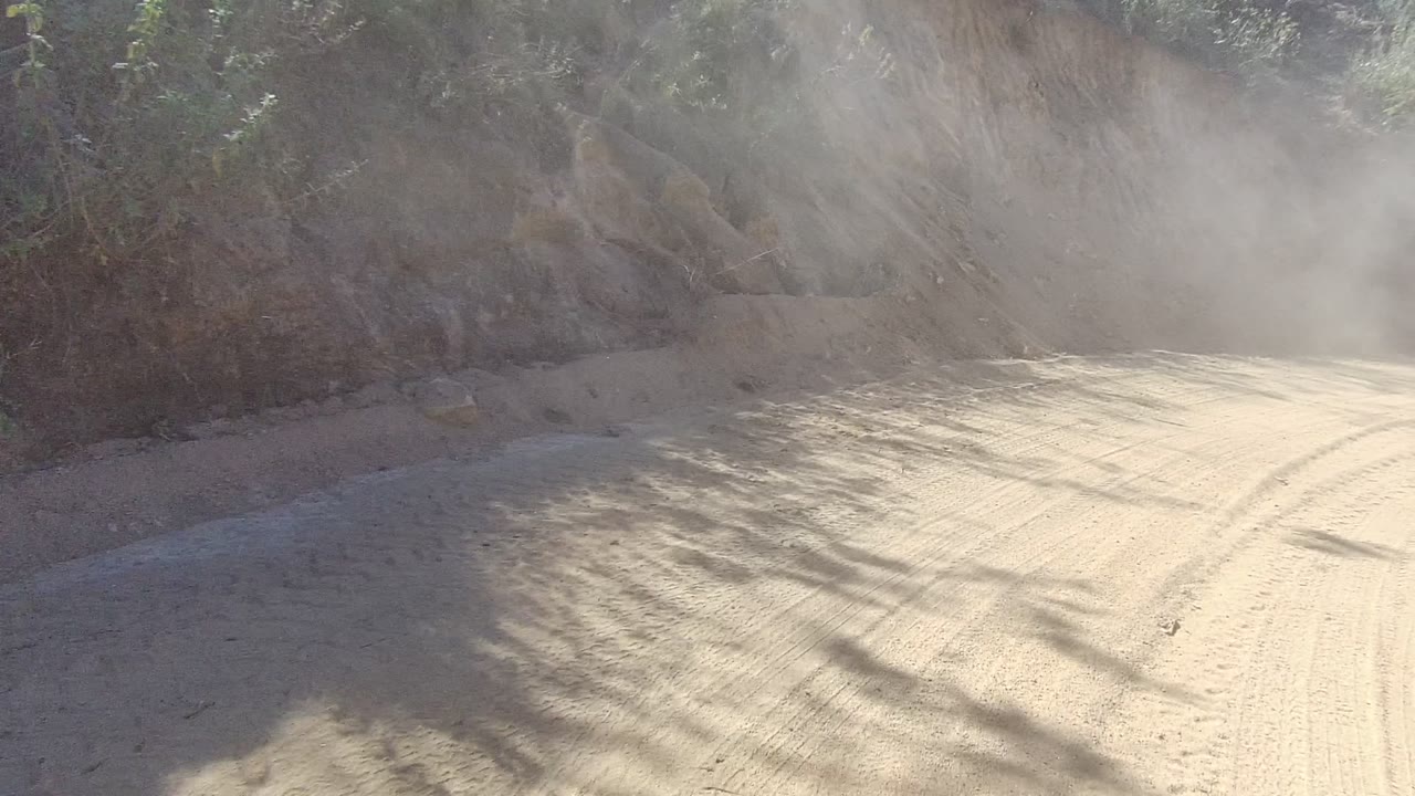 Bright sun lights a dusty bend on an unpaved Los Cabos road, with scrub vegetation along the sides, rocky edges, and no visible people.