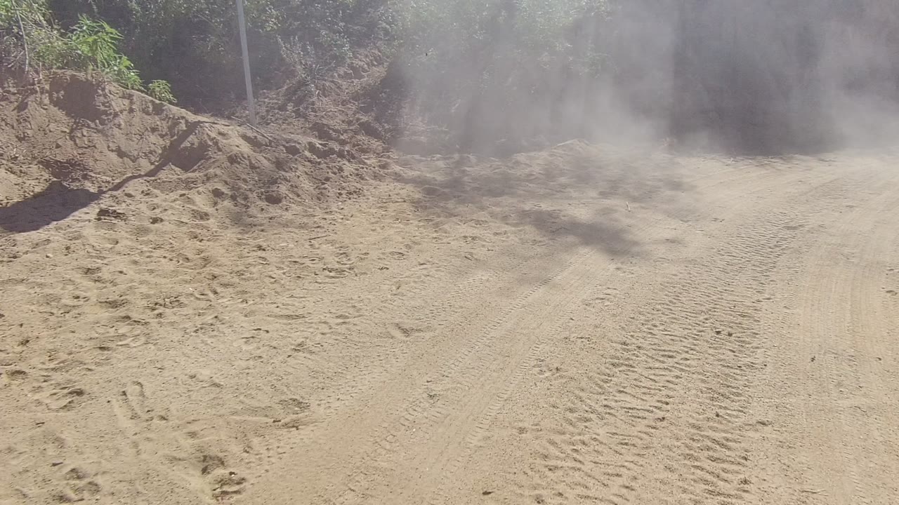 Dust rises over a sandy road bordered by dirt banks and vegetation.