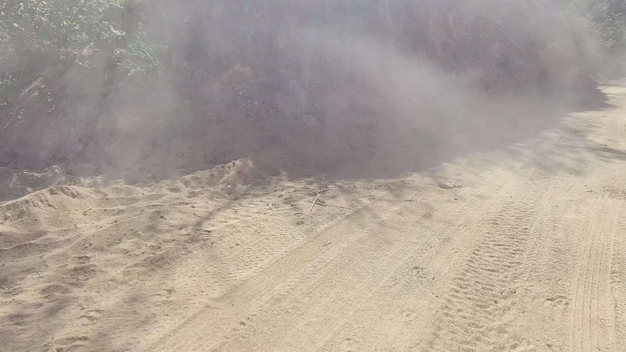 Dense dust rolls across the upper part of a Los Cabos dirt road, with rutted sand, dry banks, and roadside brush visible below, with no visible people.