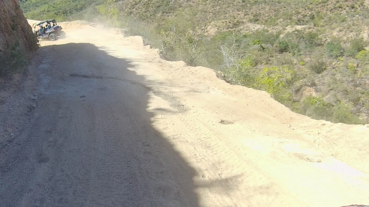 Start of a Los Cabos off-road trail view with another vehicle far ahead on the sandy road, dry hillside shadows, sparse brush, and no visible people.