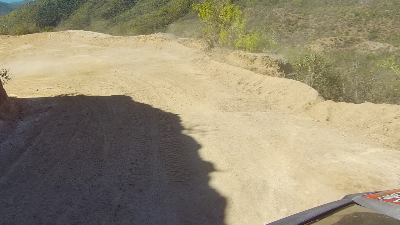 Forward vehicle view in Los Cabos with part of the hood at the bottom edge, a sandy bend, tire marks, dry brush, hillside terrain, and no visible people.