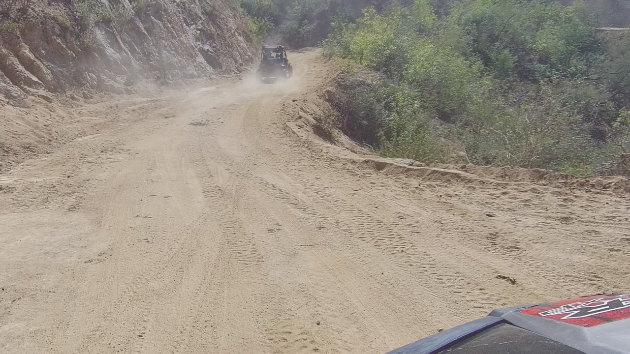 A dusty unpaved road in Los Cabos runs between rocky slopes and green brush, with a small construction vehicle in the distance and no visible people.