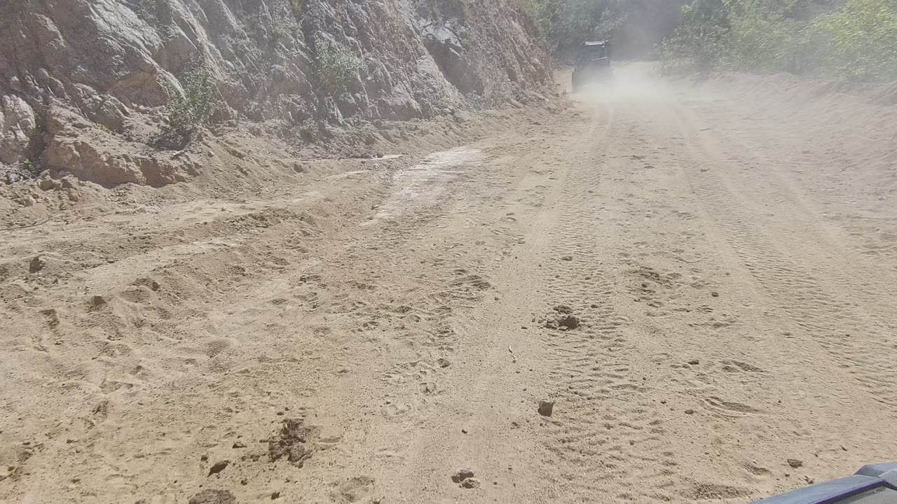 The Los Cabos dirt track curves along eroded rock and scrub vegetation, with the vehicle partly obscured by dust near the upper road and no visible people.