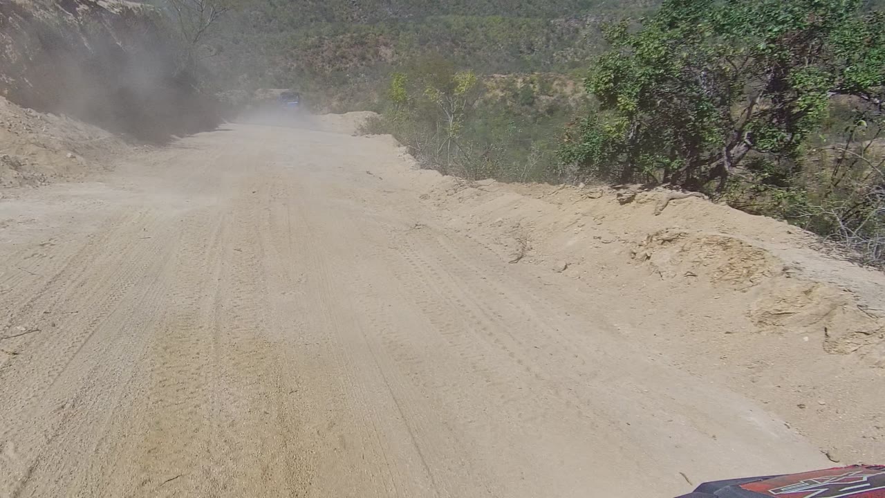 Forward view on a dusty dirt road in Los Cabos, Baja California Sur, with a sandy track, scrubby hillside, dust at left, a small vehicle edge at bottom, and no visible people.