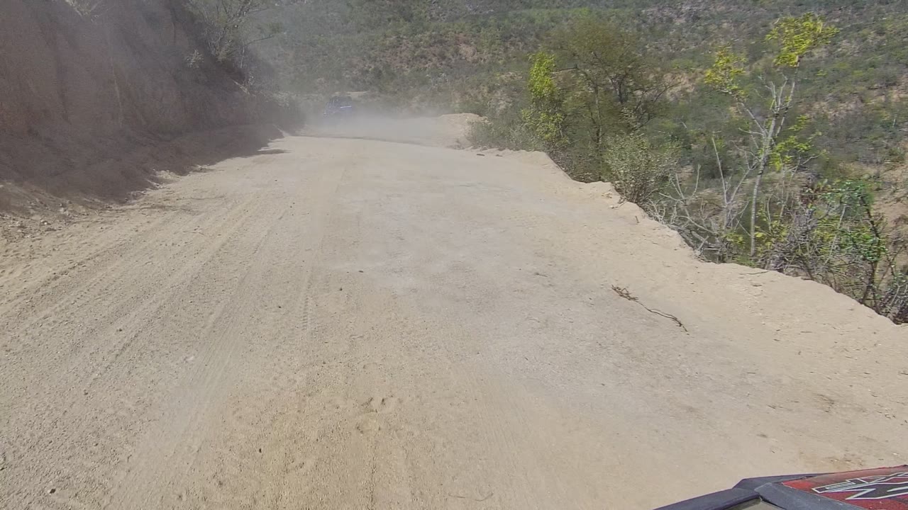 Sandy road ahead with dust in the distance and dry vegetation along the sides.