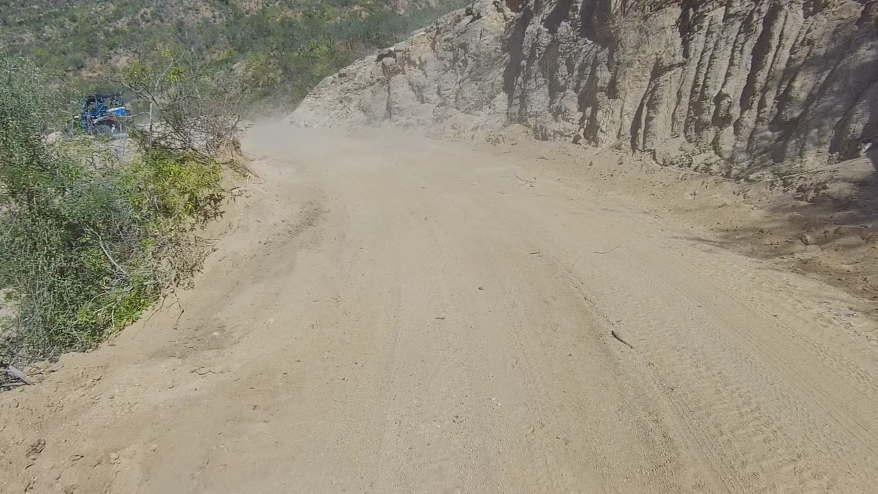 Dusty dirt road in Los Cabos, Baja California Sur, bordered by eroded rock walls and green scrub, with a small blue vehicle far left and no visible people.