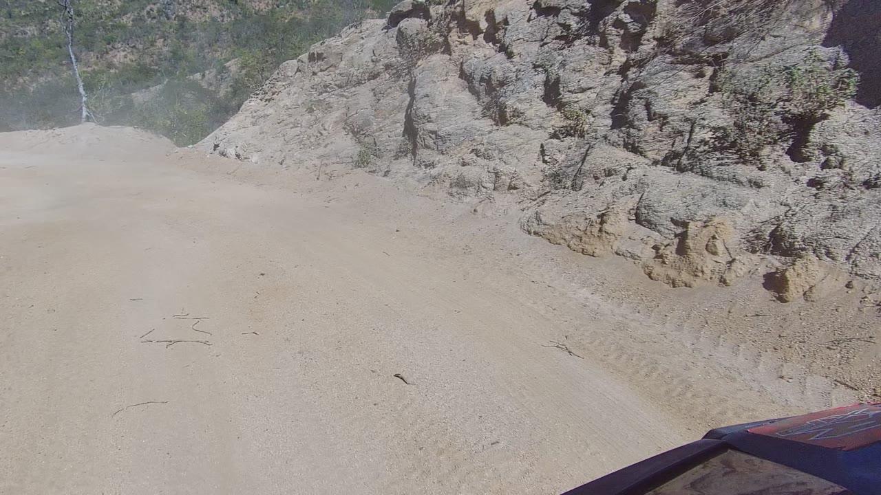 A Los Cabos dirt road runs alongside tall weathered rock formations, with pale dust drifting across the route and no visible people.