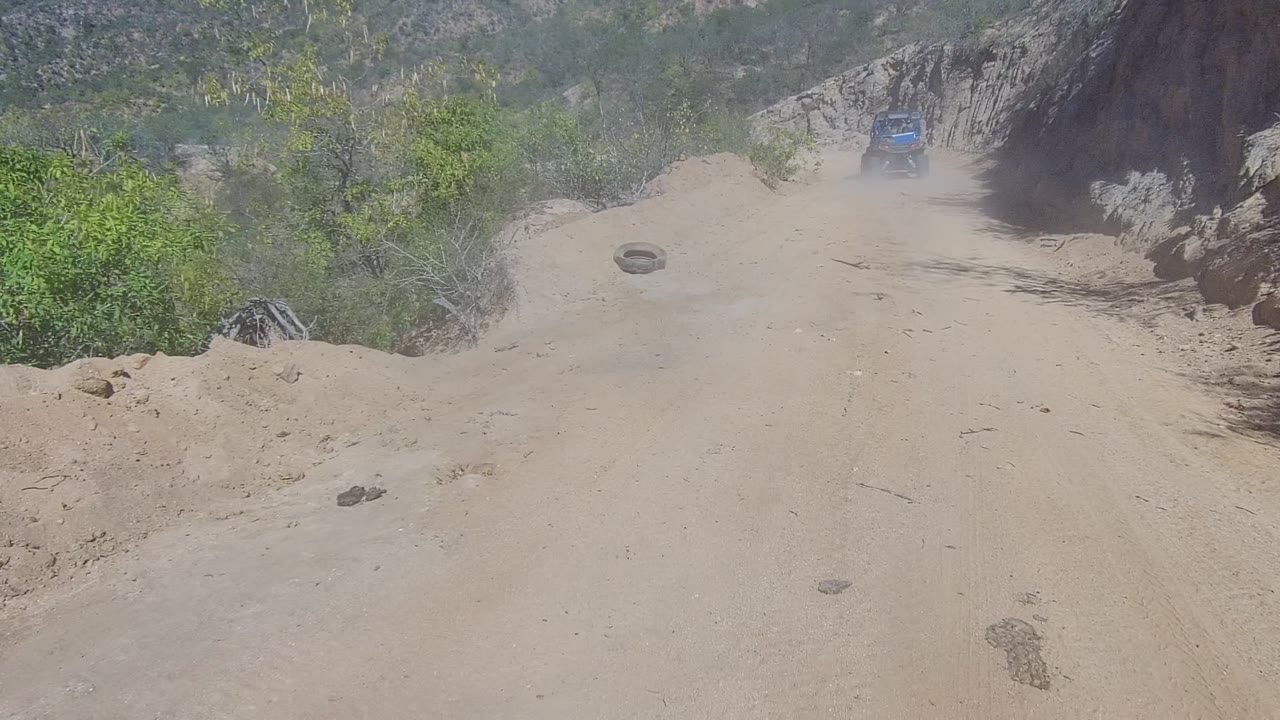 The blue truck stays distant on the right side of a sandy road bend in Los Cabos, Baja California Sur, with dust over the track, shrubs at center, and no visible people.