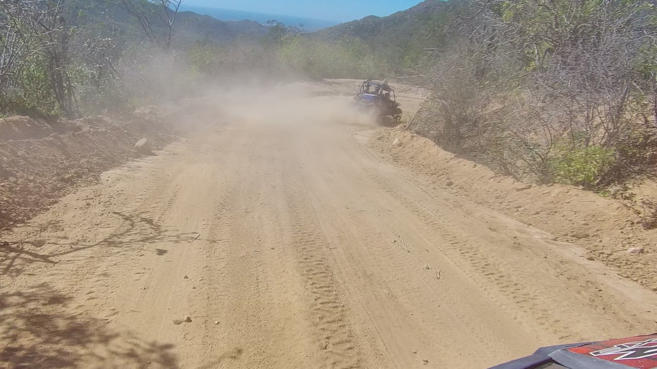 An off-road vehicle travels away on a sandy dirt road in Los Cabos, Baja California Sur, with roadside trees, low hills, and no visible people.