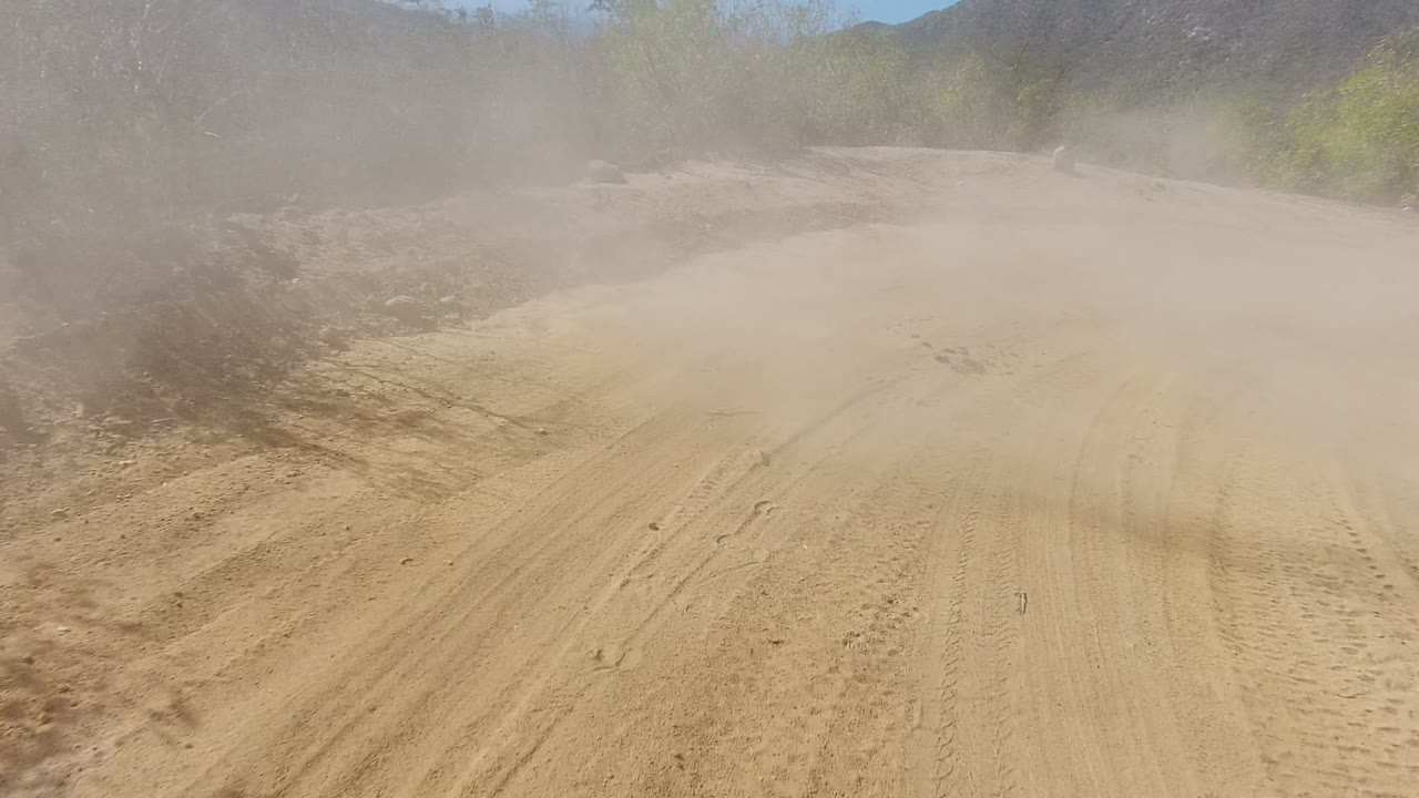 A tan dust cloud hangs over the sandy road in Los Cabos, Baja California Sur, partially hiding the path and brush while no visible people appear in the frame.